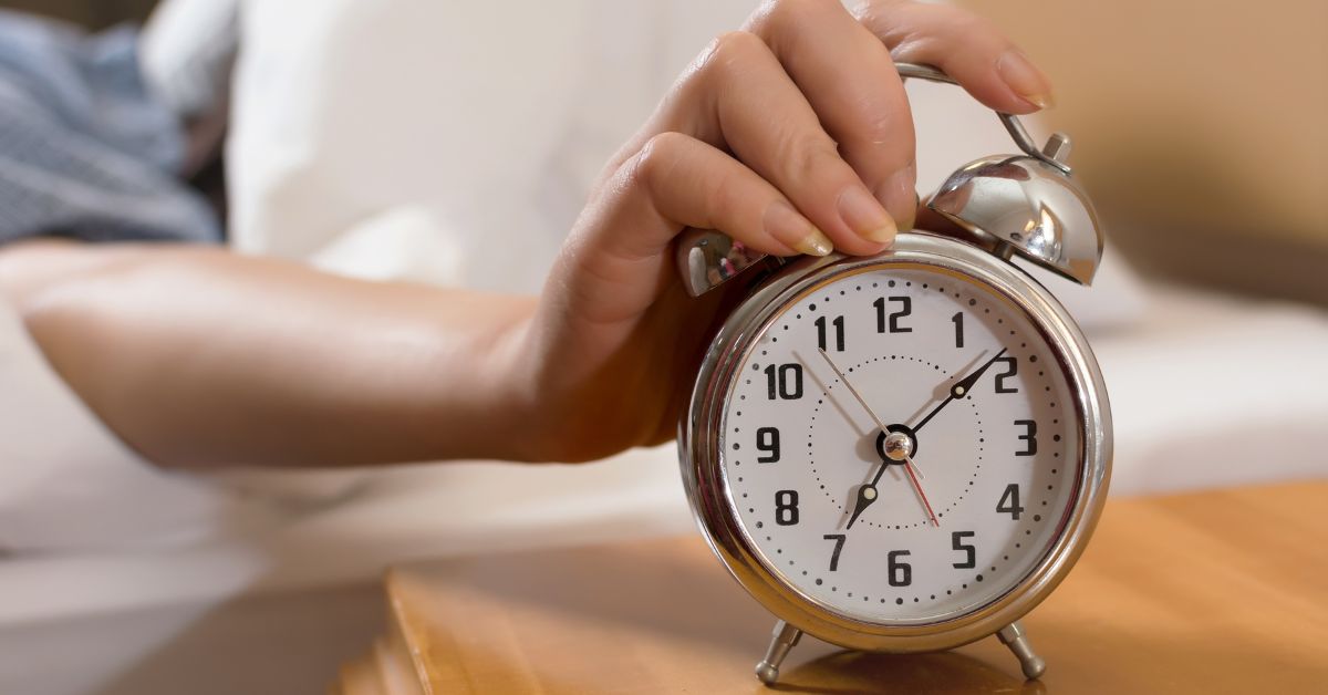 social-jetlag-alarm-clock-wake-time Hand reaching for alarm clock on bedside table, representing consistent wake time