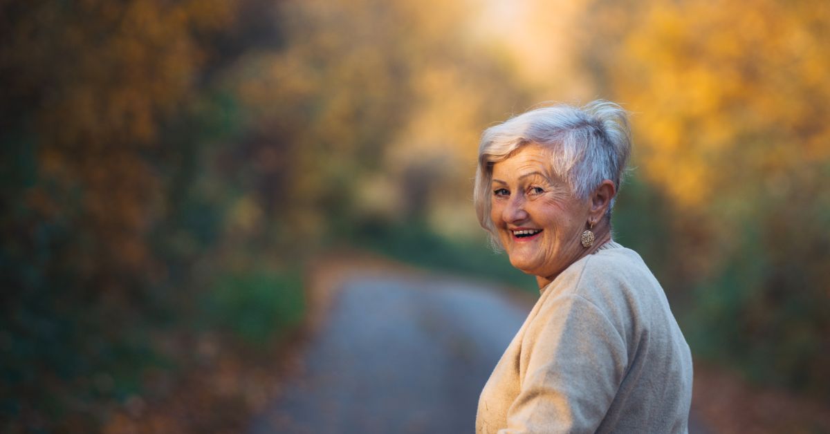 walking-outdoors-energy-stressfreelongevity Smiling older woman outdoors on a path surrounded by autumn trees