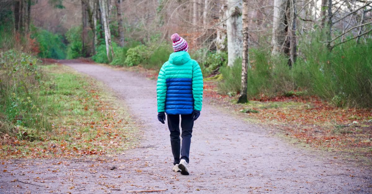 A person walking on a leaf-covered path through a wooded area in autumn, wearing a warm jacket and hat.