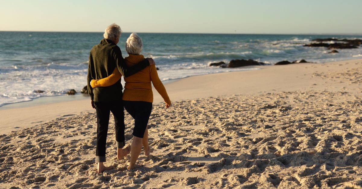 An older couple walking together along a beach at sunset, his arm around her shoulder, both looking out toward the sea.