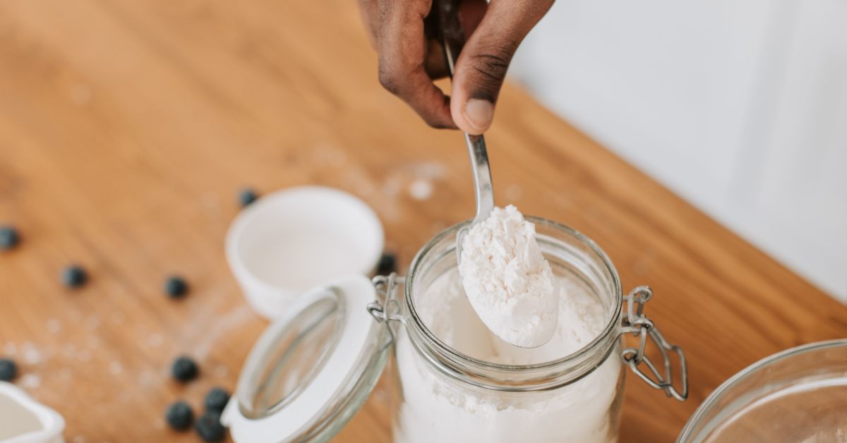 Hand scooping white creatine powder from an open glass jar on a wooden kitchen counter with blueberries scattered nearby