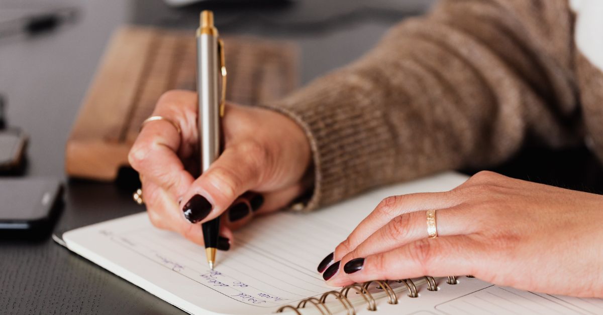 A woman writing notes in a spiral-bound journal at a desk, with a pen in her hand and a laptop visible beside her.