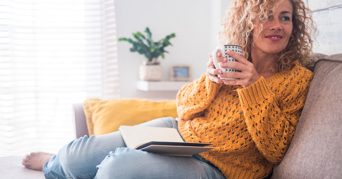 A woman in her 40s sitting on a sofa at home, holding a mug of tea with a book resting in her lap, in a quiet moment of reflection.