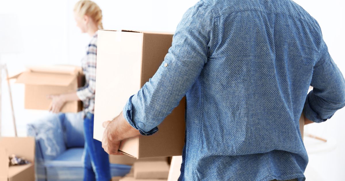 A man carries a cardboard box while a woman packs another in the background