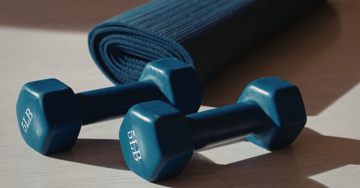 A pair of light blue 5lb dumbbells on a wooden floor with a rolled yoga mat behind them
