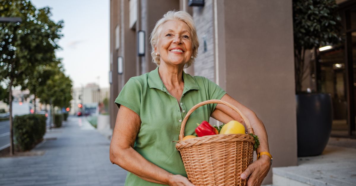 Smiling woman in her 60s carrying a basket of fresh produce one-handed — illustrating muscle as everyday capability after 40