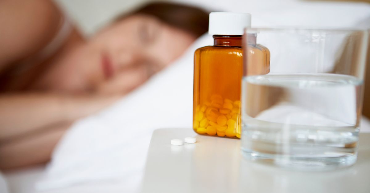 safest-sleep-aid-bedside-pills-stressfreelongevity Pill bottle and glass of water on a bedside table with a person sleeping in the background