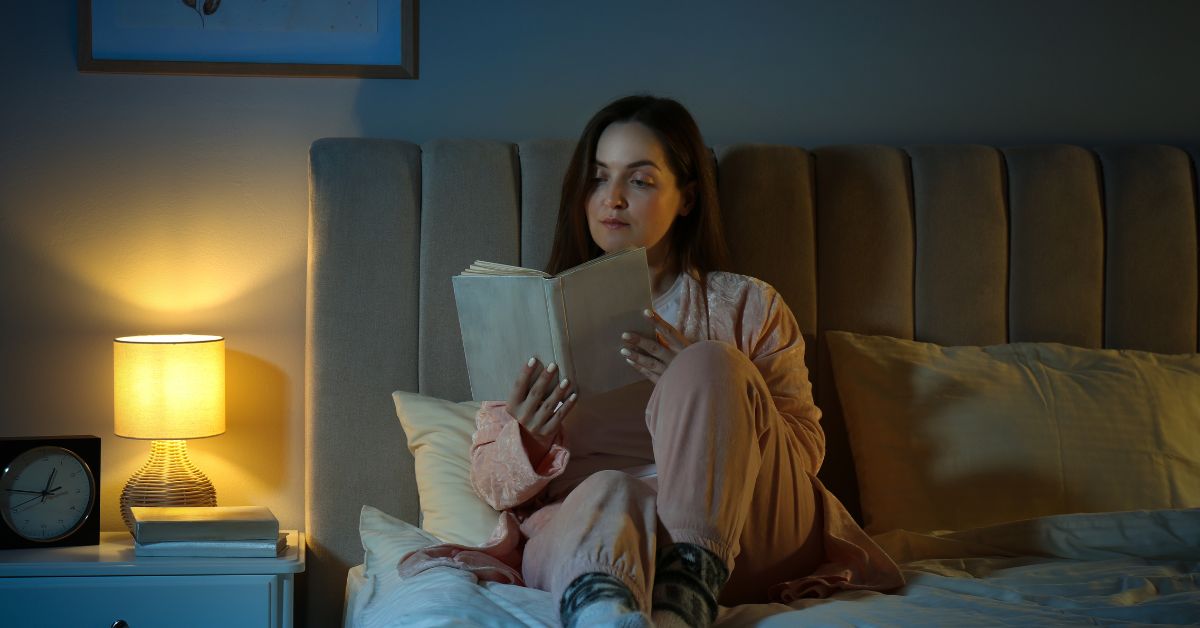 safest-sleep-aid-reading-before-bed-stressfreelongevity Woman reading a book in bed with a warm bedside lamp
