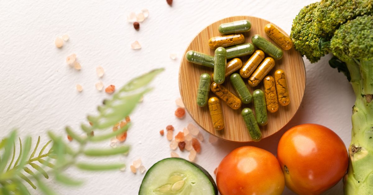 safest-sleep-aid-supplements-natural-stressfreelongevity Herbal supplement capsules in a wooden bowl surrounded by broccoli, tomatoes, and cucumber