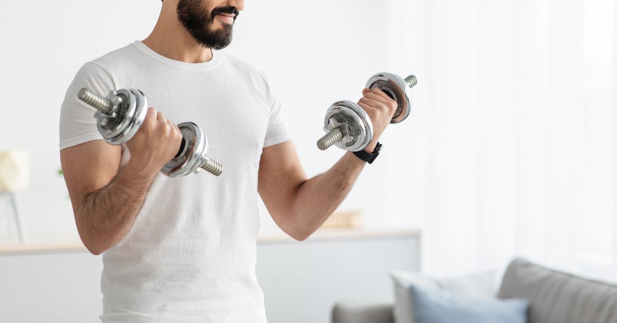 how-much-strength-training-per-week-home-dumbbell-stressfreelongevity Man doing a strength training session at home with dumbbells in a bright living room