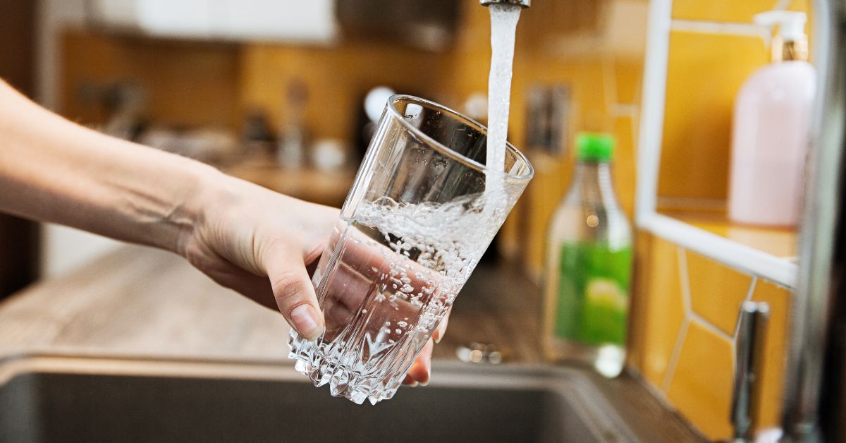 reduce-pfas-exposure-forever-chemicals-stressfreelongevity Person filling a glass of water from a kitchen tap, illustrating how to reduce PFAS exposure from forever chemicals in drinking water