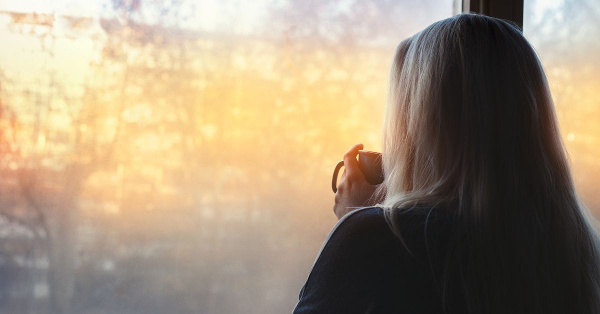 Building sleep consistency — a person at a sunlit window with morning coffee, starting the day at a regular time