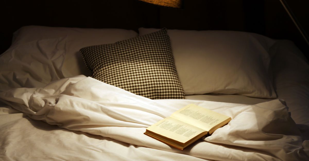 8-hour-sleep-myth-evening-wind-down-stressfreelongevity An open book on a bed under warm lamplight, a calm screen-free wind-down.