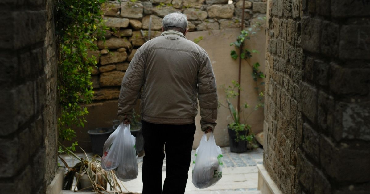 grip-strength-independence-older-man-carrying-shopping-stressfreelongevity Older man carrying shopping bags home through a stone courtyard