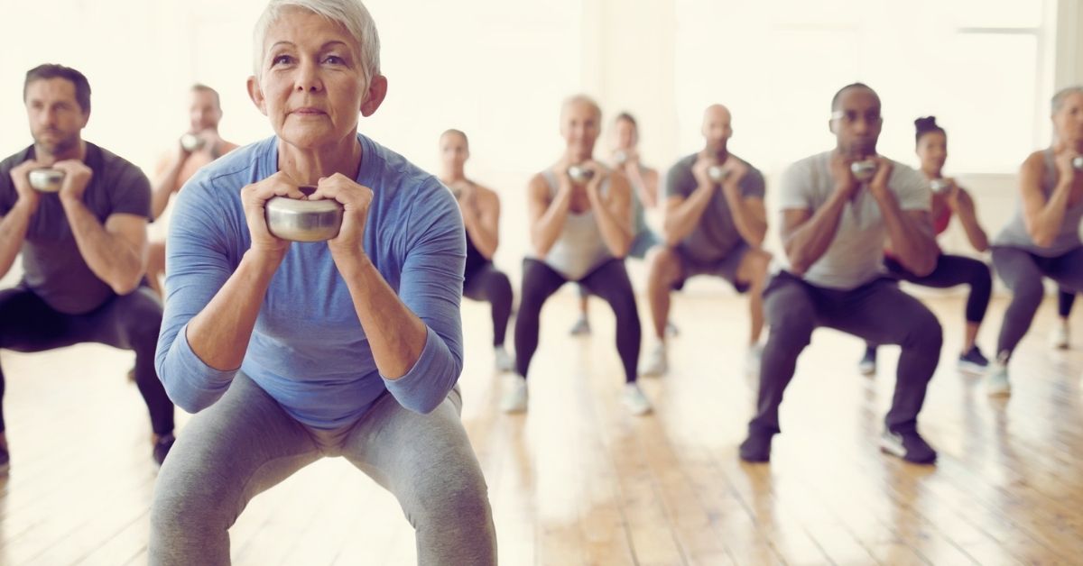 Older woman squatting with a kettlebell in a group fitness class, building the leg strength measured by the sitting-rising test